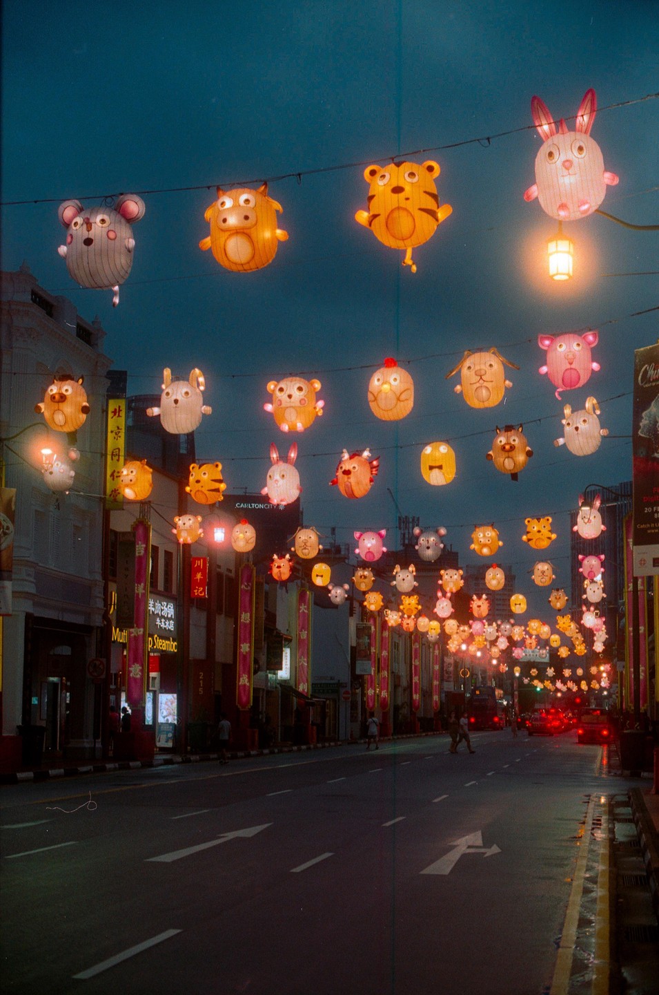 Animal-shaped lanterns hang above a street with buildings and cars at dusk.