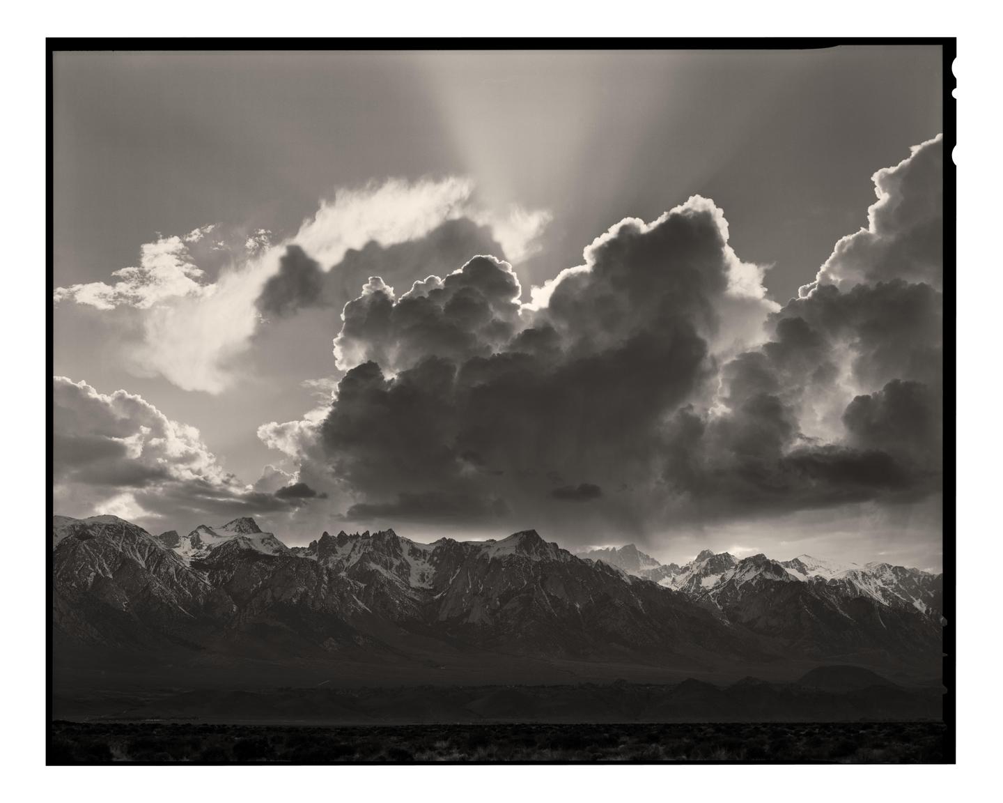 Sun rays shine through dramatic clouds over a mountain range with snow-capped peaks.