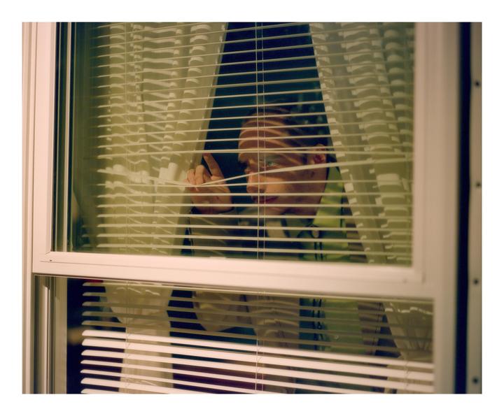 A man peeks through window blinds, with light stripes across his face.