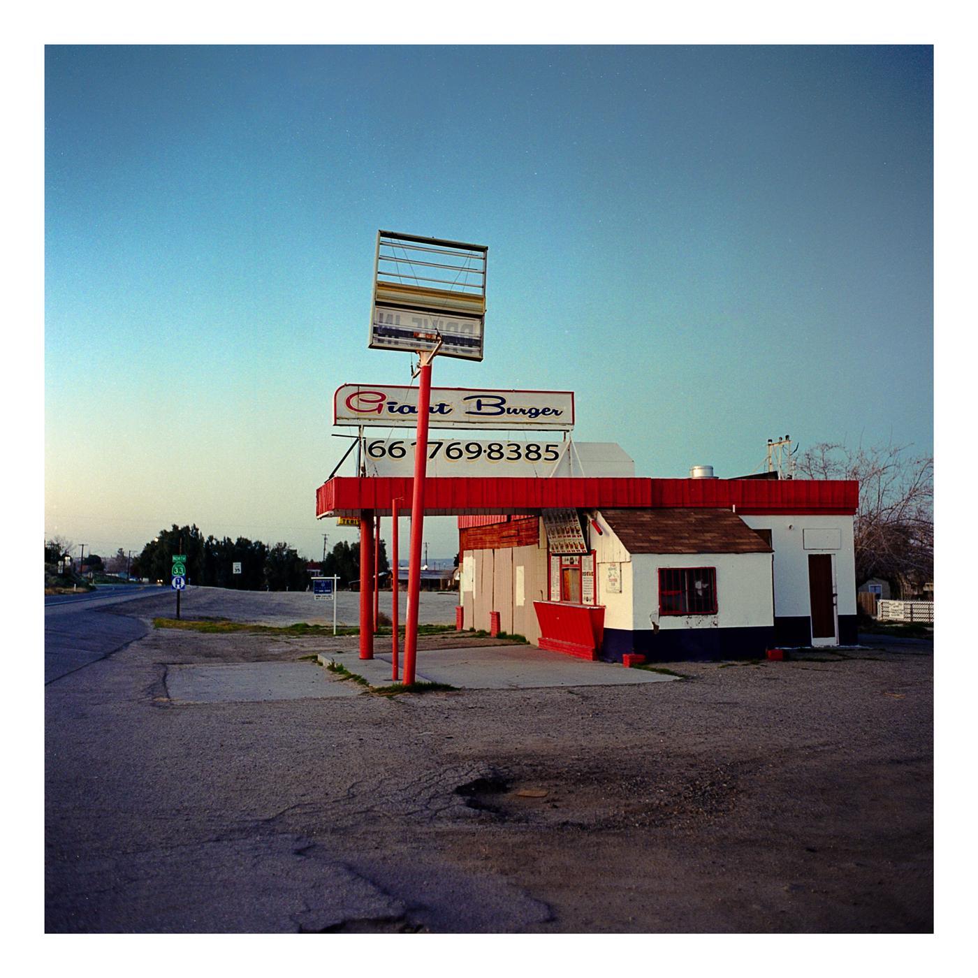 An abandoned Giant Burger stand with a large sign stands beside a road under a clear sky.