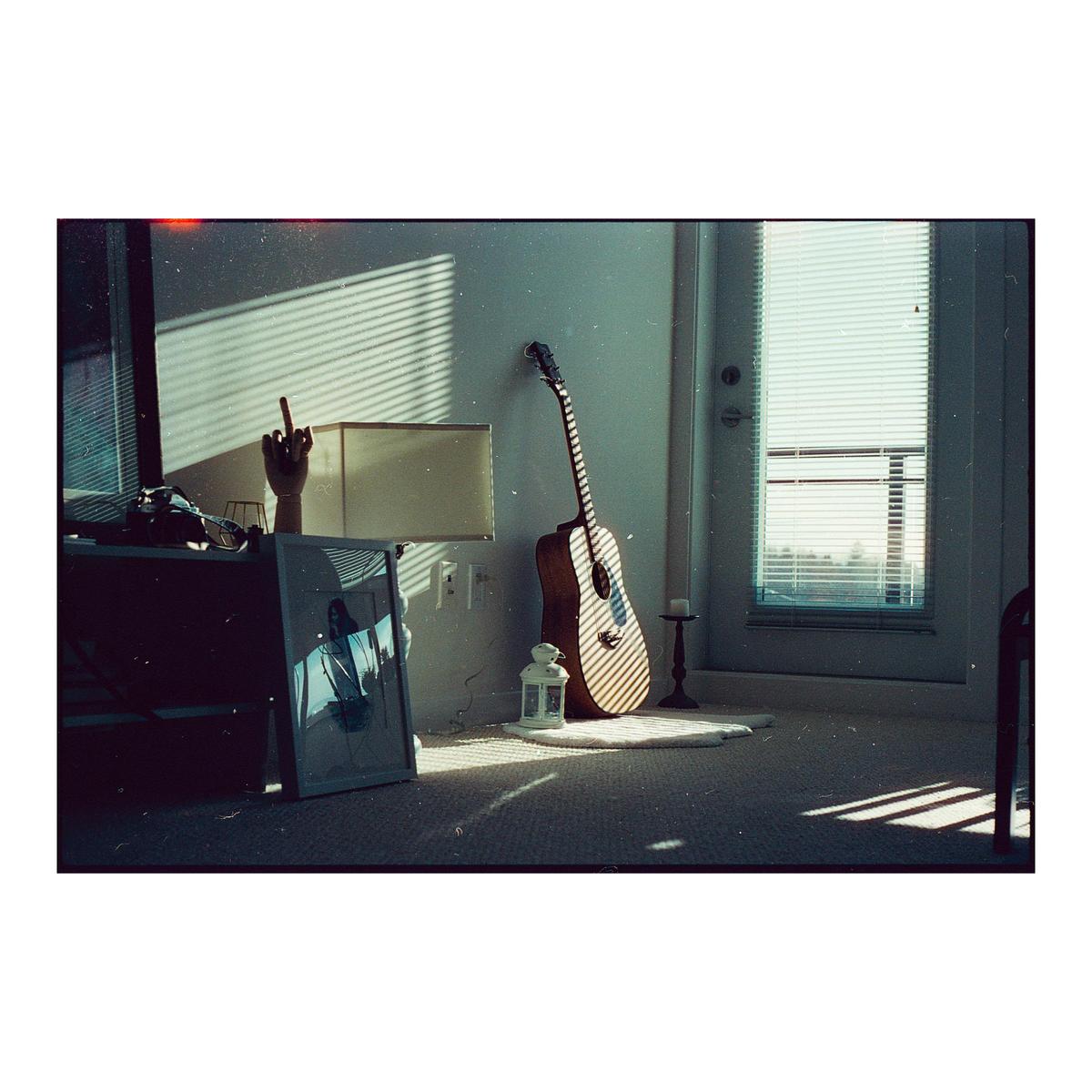 Sunlight streams through blinds onto a guitar, lamp, and framed picture in a room.