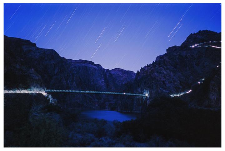 A long exposure shot of a suspension bridge over a body of water in a canyon at night, with star trails in the sky.
