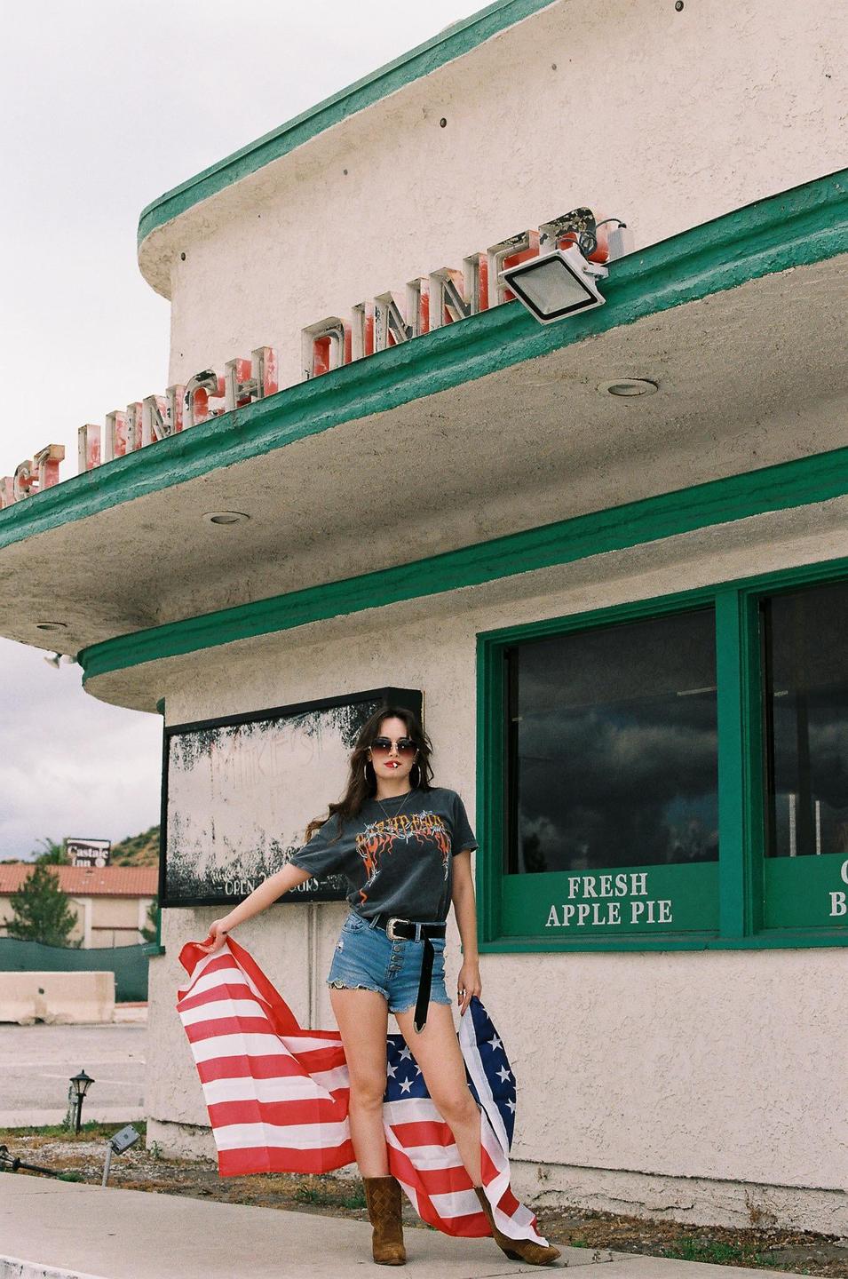 A woman stands in front of a diner holding an American flag.