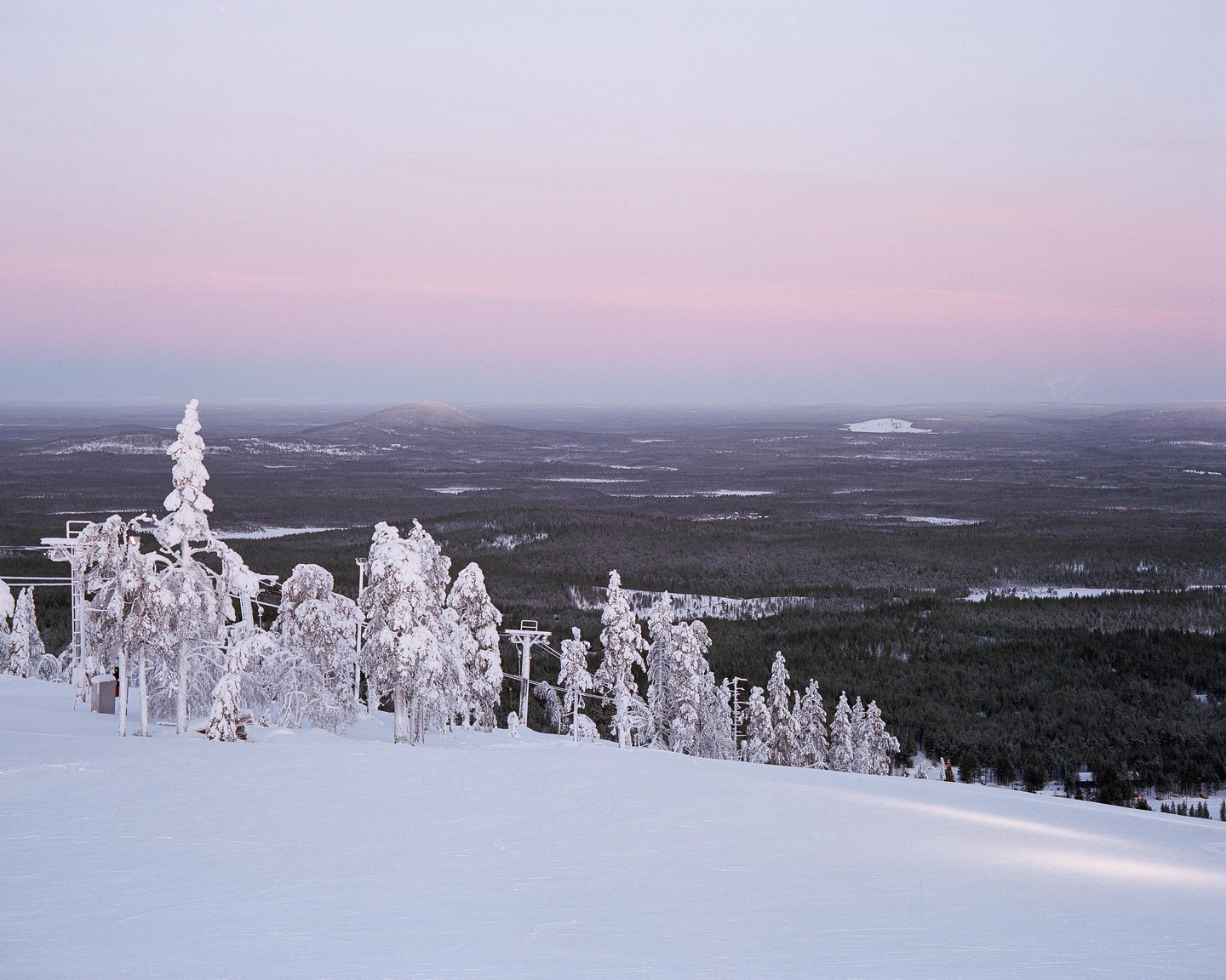 Snow-covered trees and a ski lift are visible on a mountain slope with a vast forest and hills in the distance under a pastel sky.