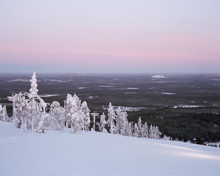 Snow-covered trees and a ski lift are visible on a mountain slope with a vast forest and hills in the distance under a pastel sky.