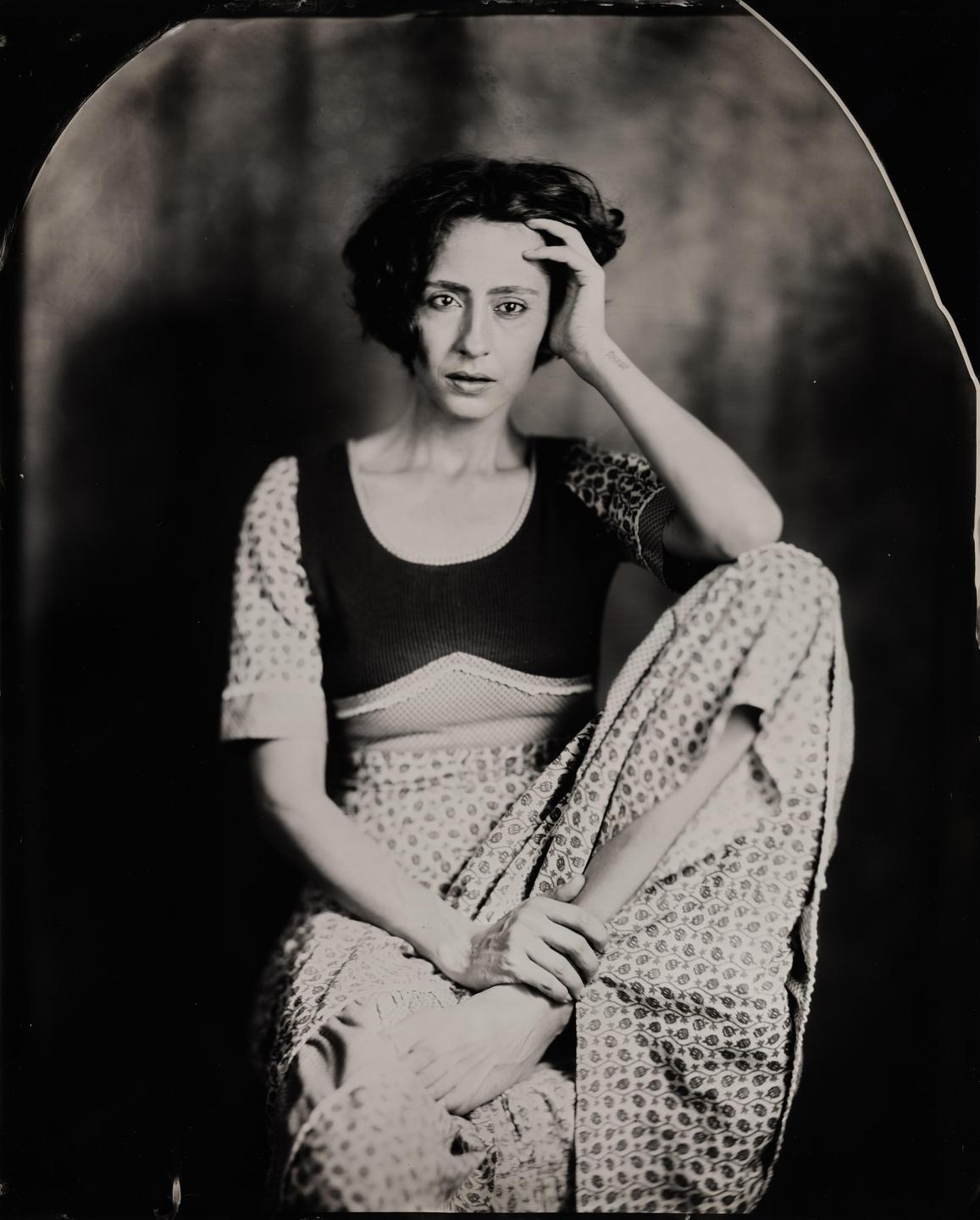 A woman with short, dark hair sits with one leg bent and her hand resting on her head.