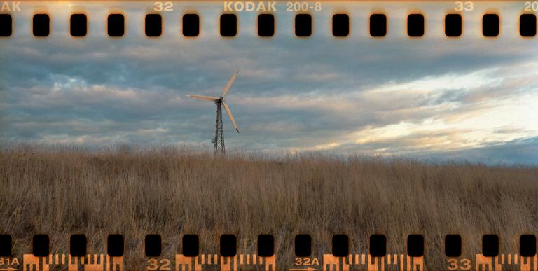 A wind turbine stands in a field of tall grass under a cloudy sky.