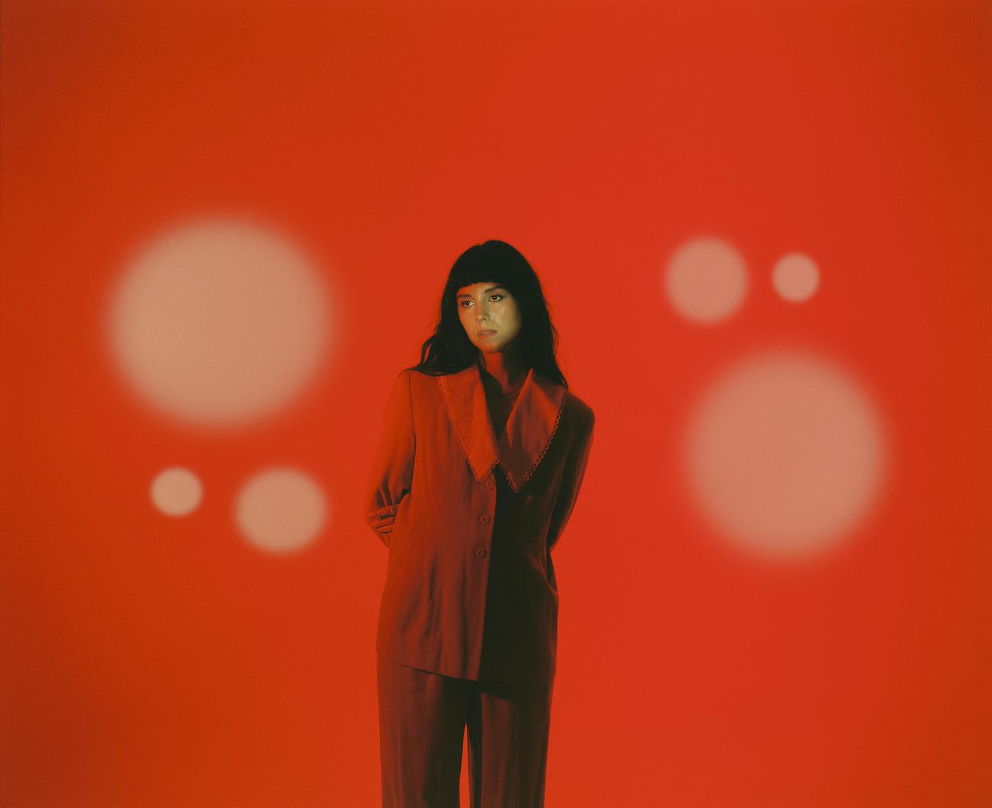 A woman in a suit stands against a red background with several white circles.