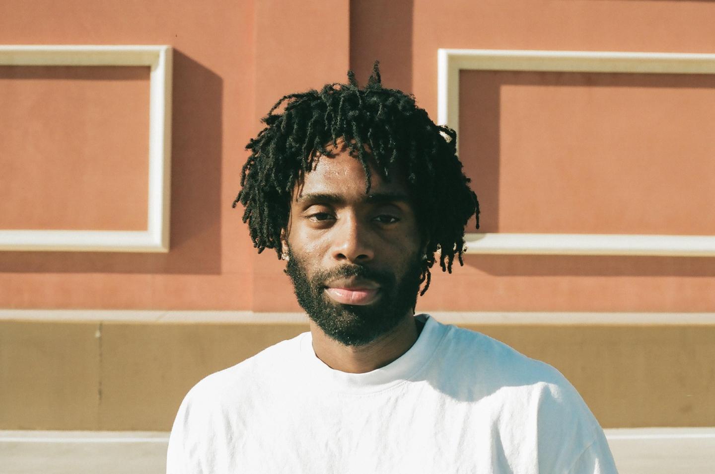 A man with dreadlocks and a beard looks directly at the viewer against a peach-colored wall with white trim.