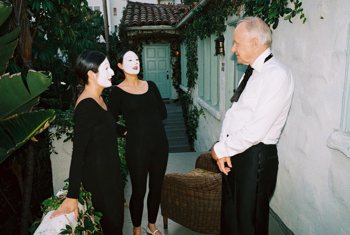 Two women in mime makeup and black outfits stand with a man in a white shirt and black tie outside a building with green trim.