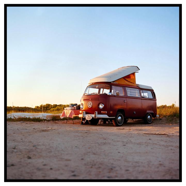 A red camper van with its top popped up is parked on a dirt road next to a small table with a red and white checkered tablecloth.