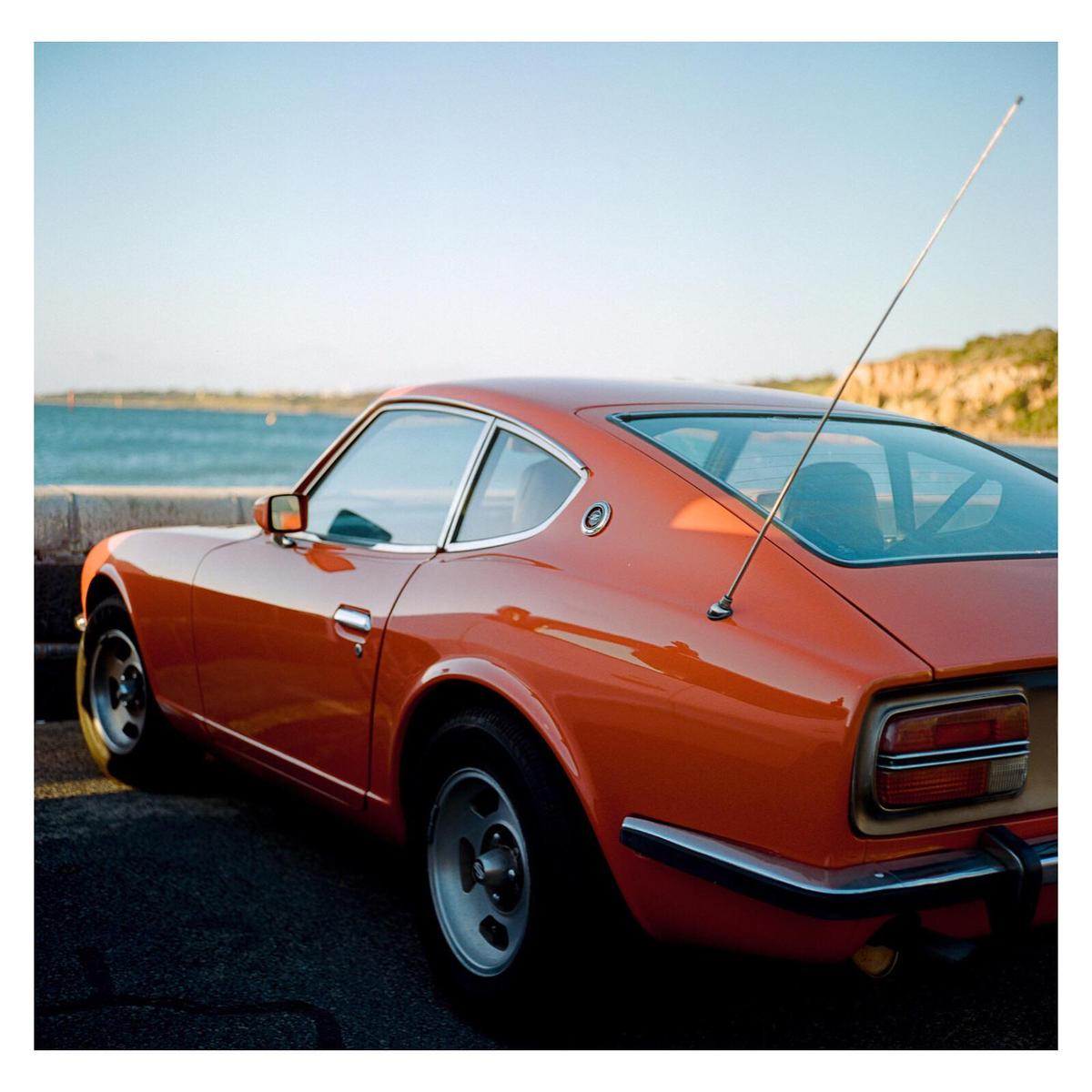 An orange sports car is parked on a paved surface next to a body of water with a distant shoreline.