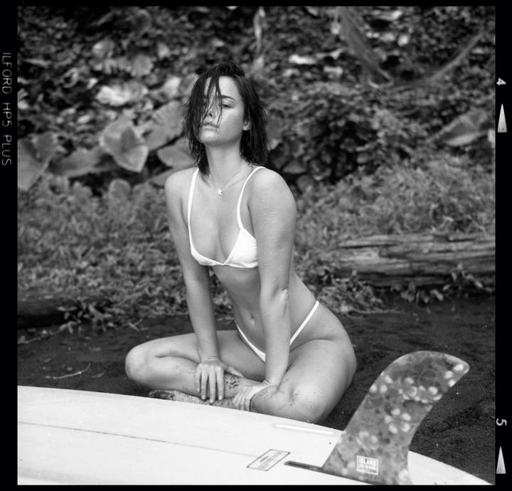 A woman in a bikini sits on the ground next to a surfboard.