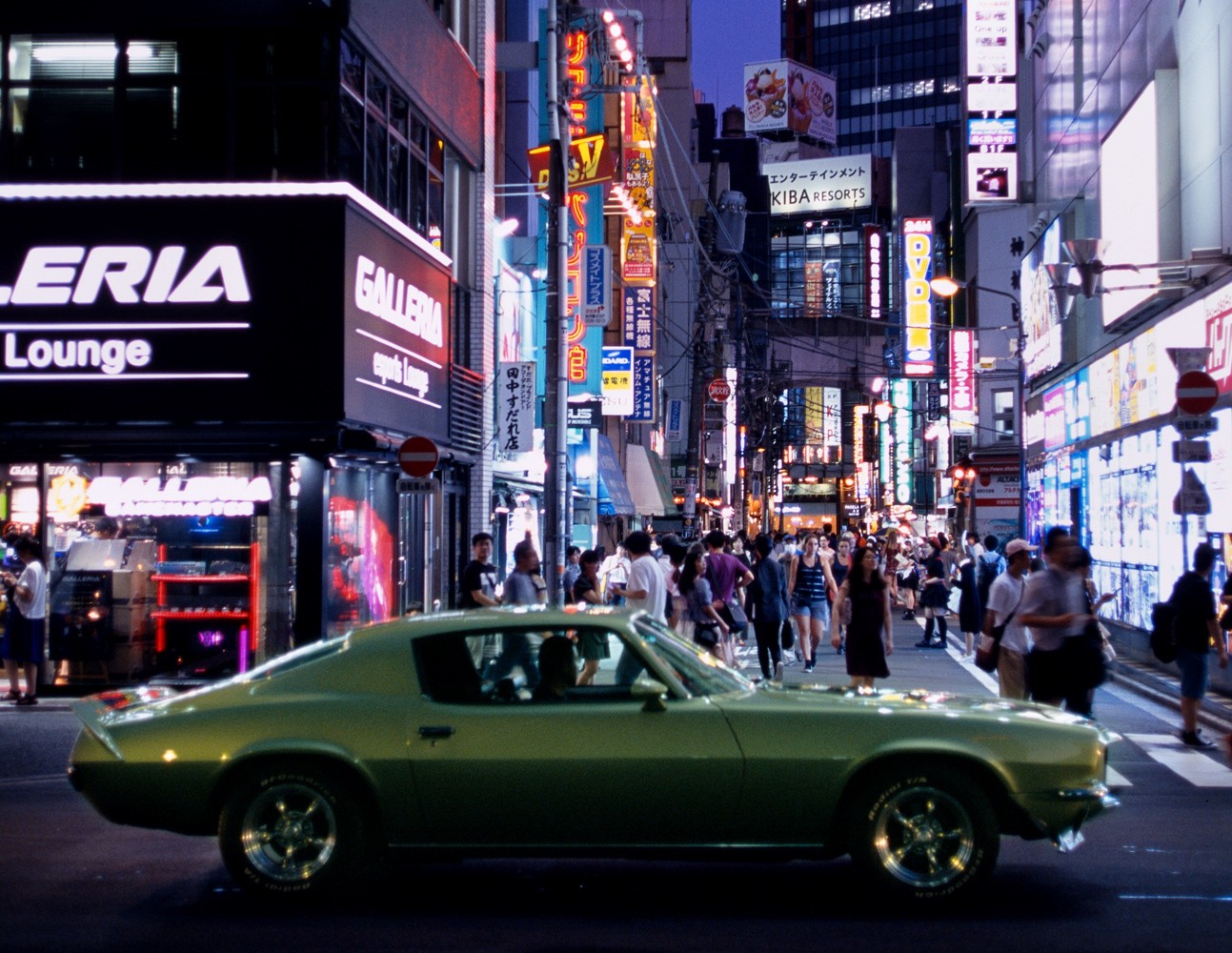 A green muscle car drives through a busy street at night, surrounded by neon signs and pedestrians.
