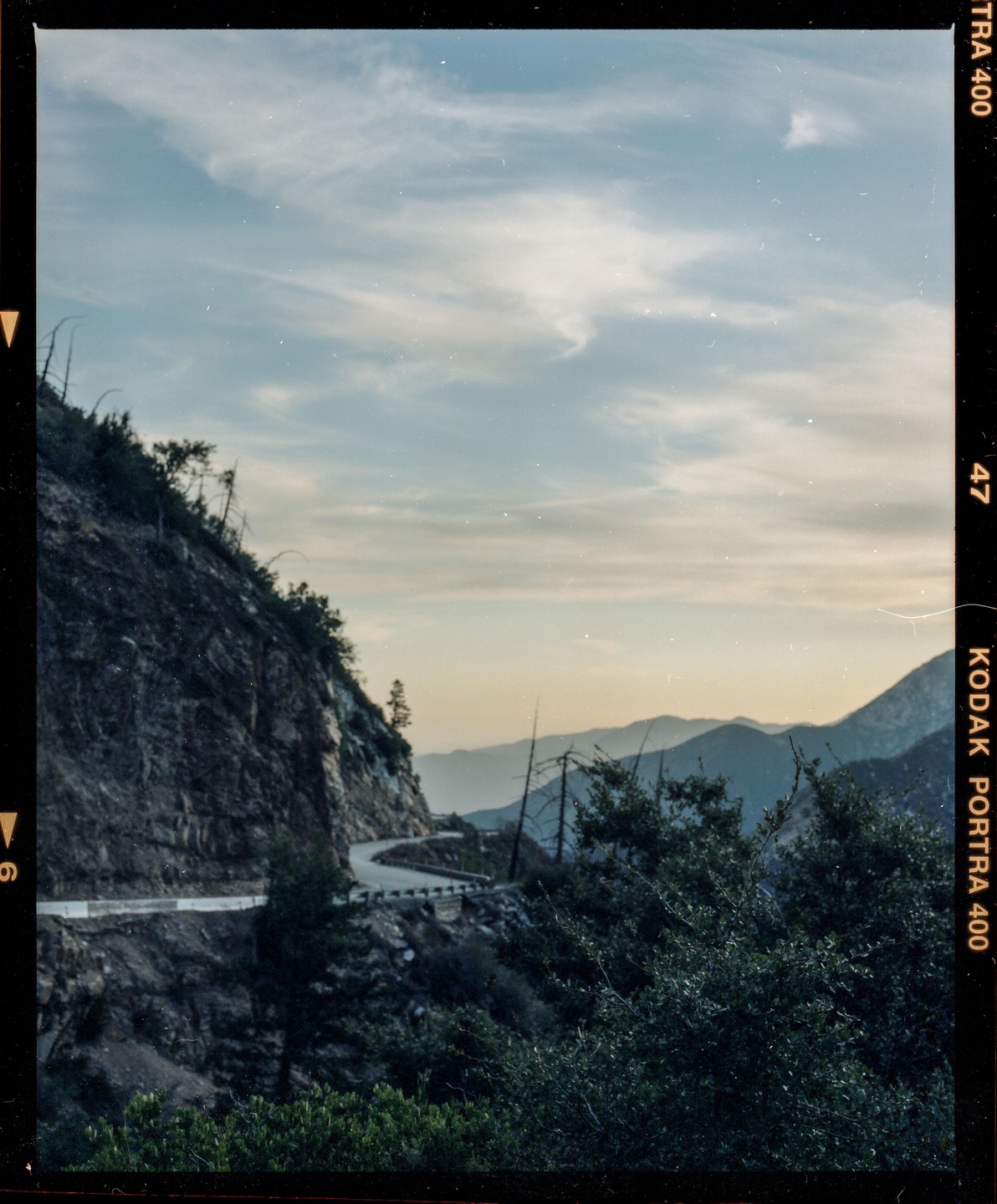 A winding mountain road is visible on the side of a steep, rocky mountain, with distant mountains and a cloudy sky in the background.