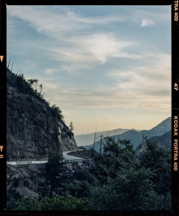 A winding mountain road is visible on the side of a steep, rocky mountain, with distant mountains and a cloudy sky in the background.