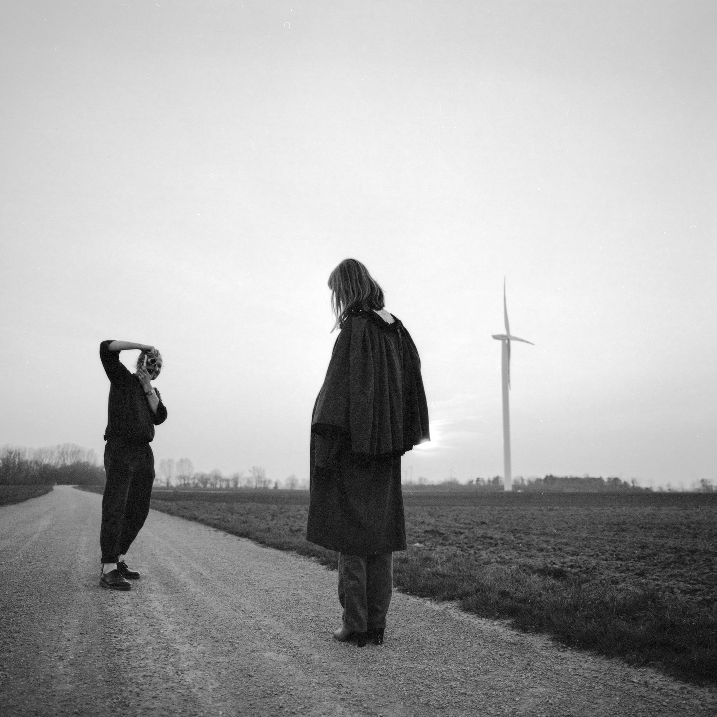 A person in a long coat stands on a country lane facing away from another person wearing a mask, with a wind turbine in the background.