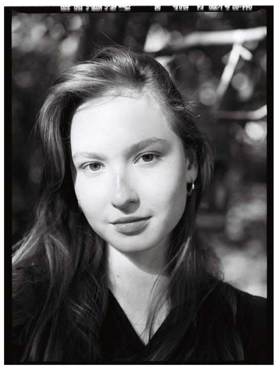 A young woman with long hair looks directly at the viewer in a black and white portrait.