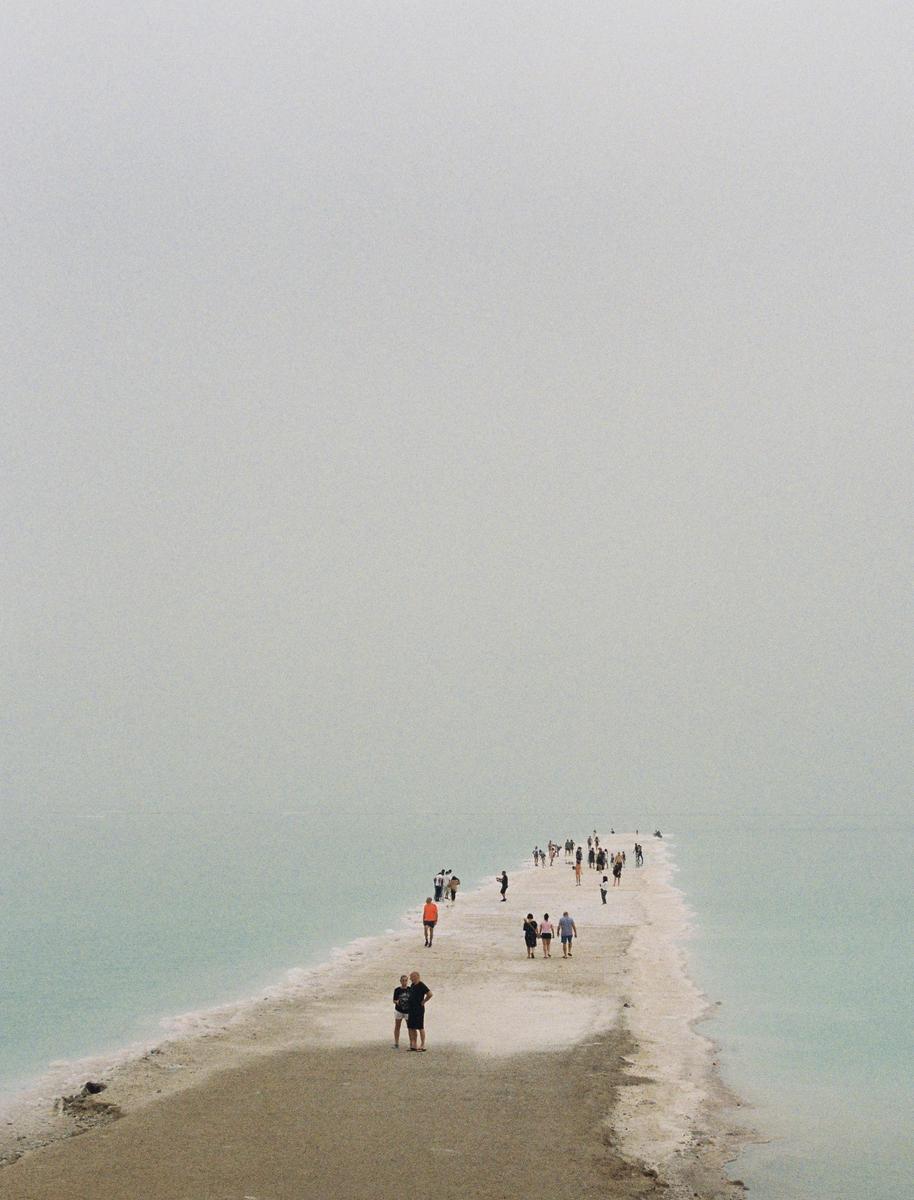 A group of people walk along a narrow strip of land extending into the hazy Dead Sea.