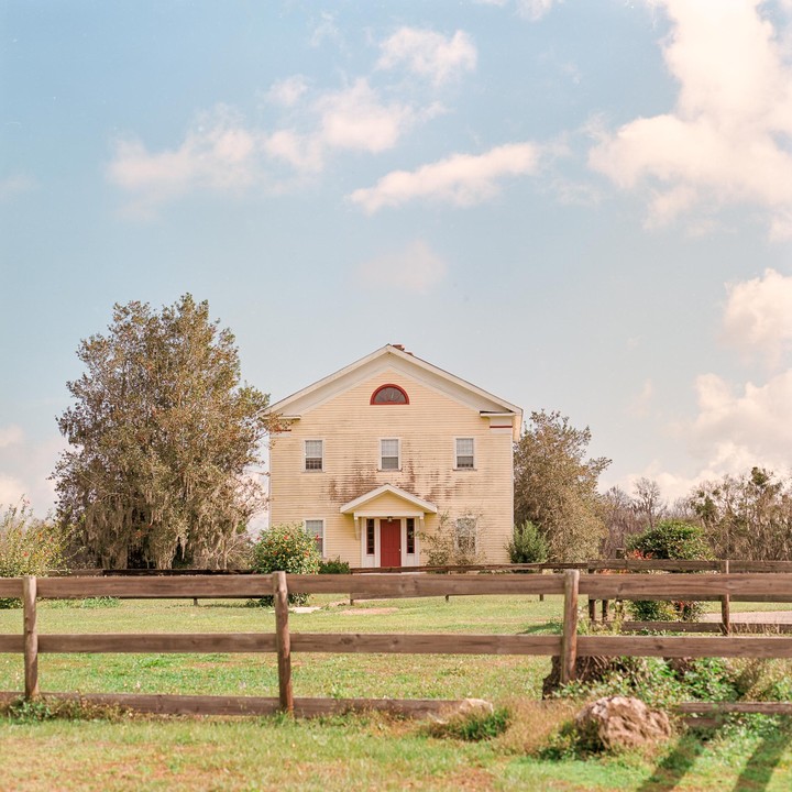 A yellow house with a red door stands behind a wooden fence in a grassy field under a cloudy sky.