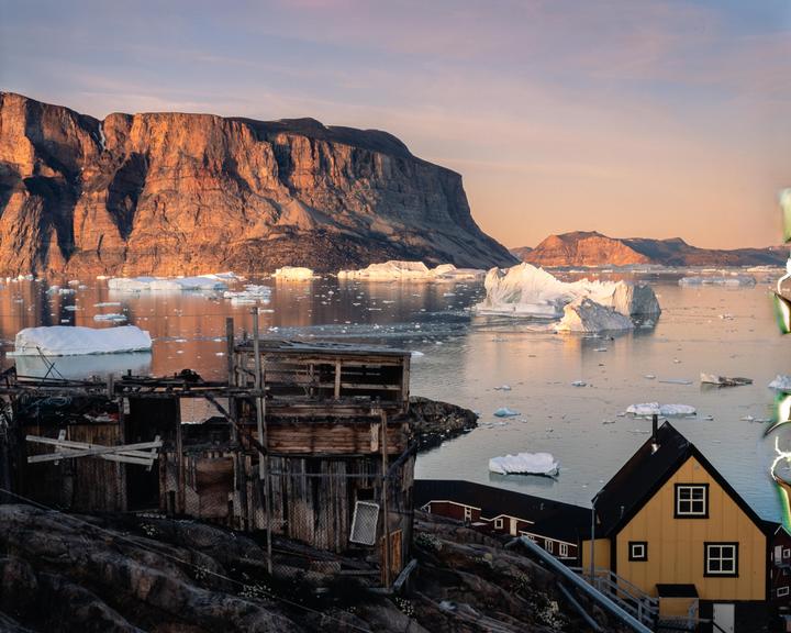 A small settlement with wooden houses sits on a rocky shore, facing a body of water filled with icebergs and backed by a large mountain range under a golden hour sky.