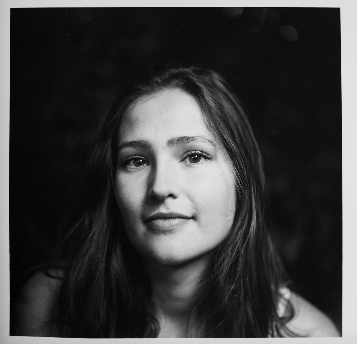 A woman with long hair looks directly at the viewer in a black and white portrait.