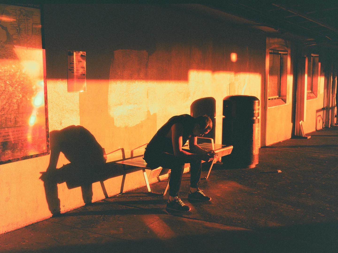 A man sits on a bench in a dark area with a bright orange light illuminating the wall behind him.
