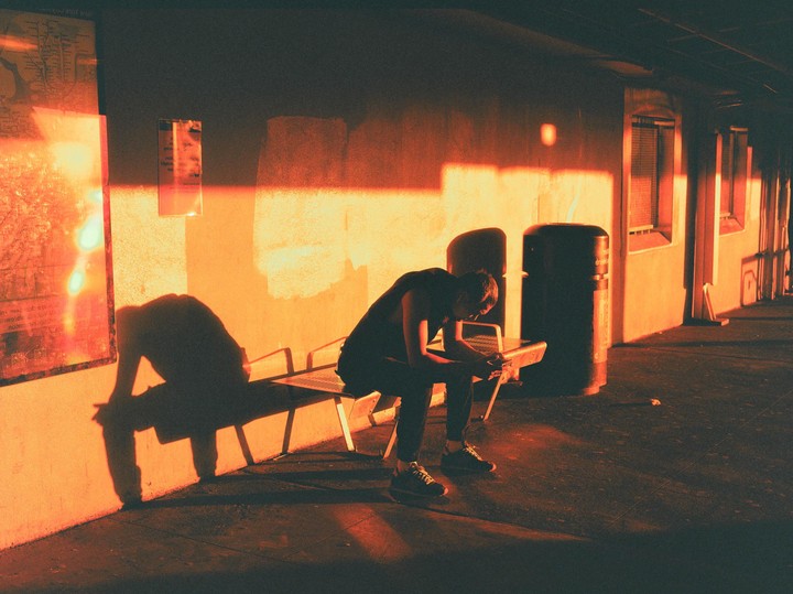 A man sits on a bench in a dark area with a bright orange light illuminating the wall behind him.