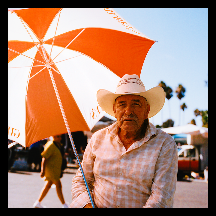 A man in a cowboy hat sits under an orange and white umbrella on a sunny day.