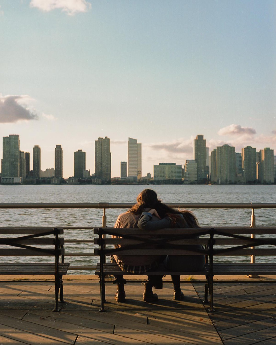 A couple sits on a bench by the water, looking at a city skyline across the river.