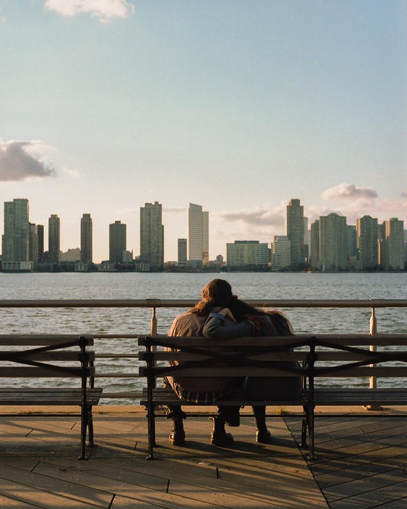 A couple sits on a bench by the water, looking at a city skyline across the river.