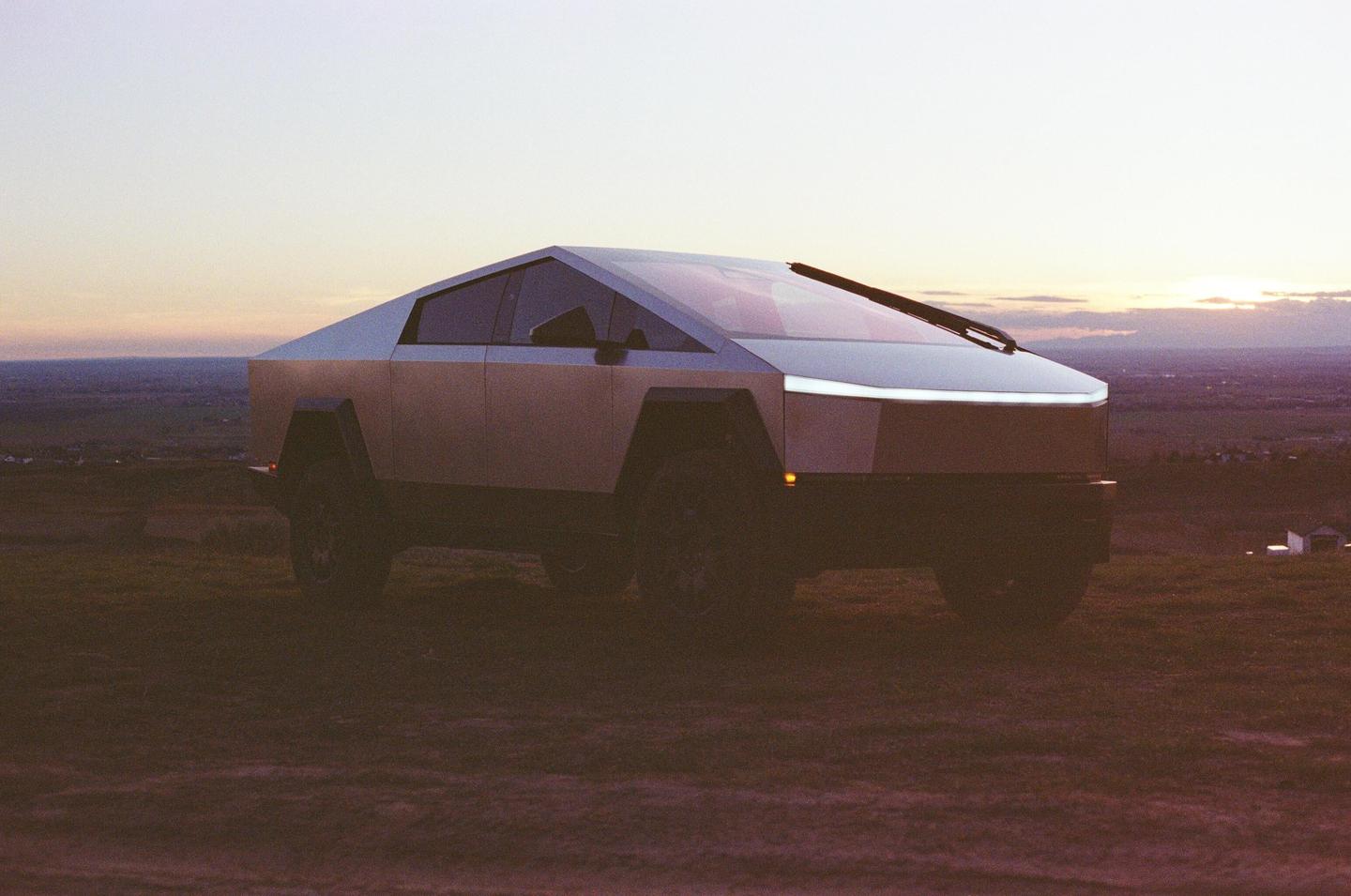 A Cybertruck is parked on a grassy hill overlooking a valley at sunset.