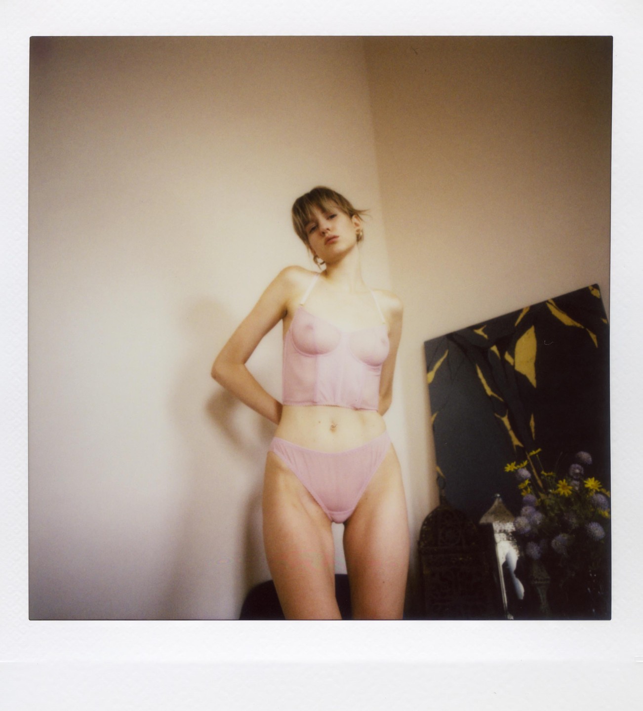 A woman in pink lingerie stands in a room with a mirror and flowers.