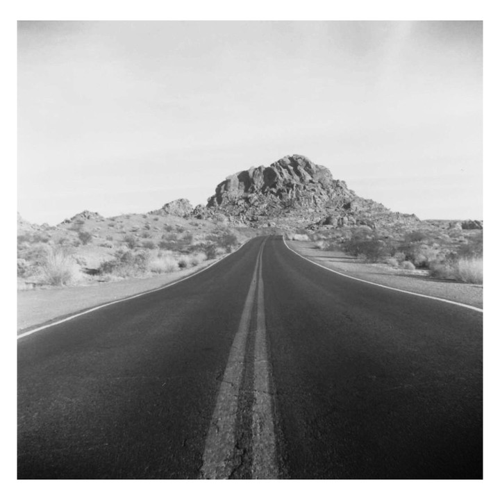 A long, empty road stretches towards a rocky mountain in a desert landscape.