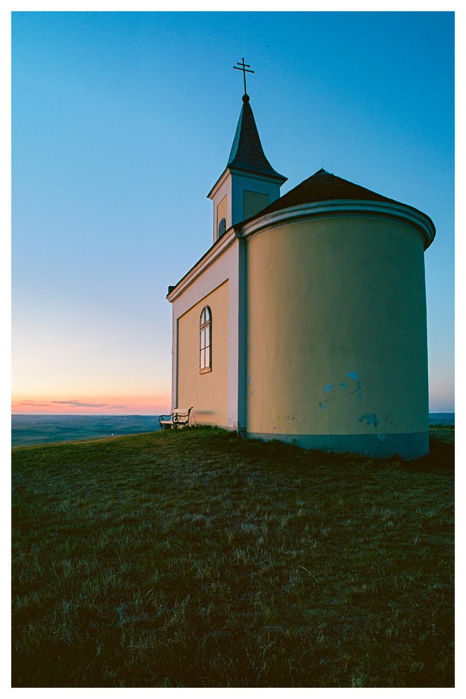 A small church with a steeple and cross stands on a grassy hill overlooking a valley at dusk.