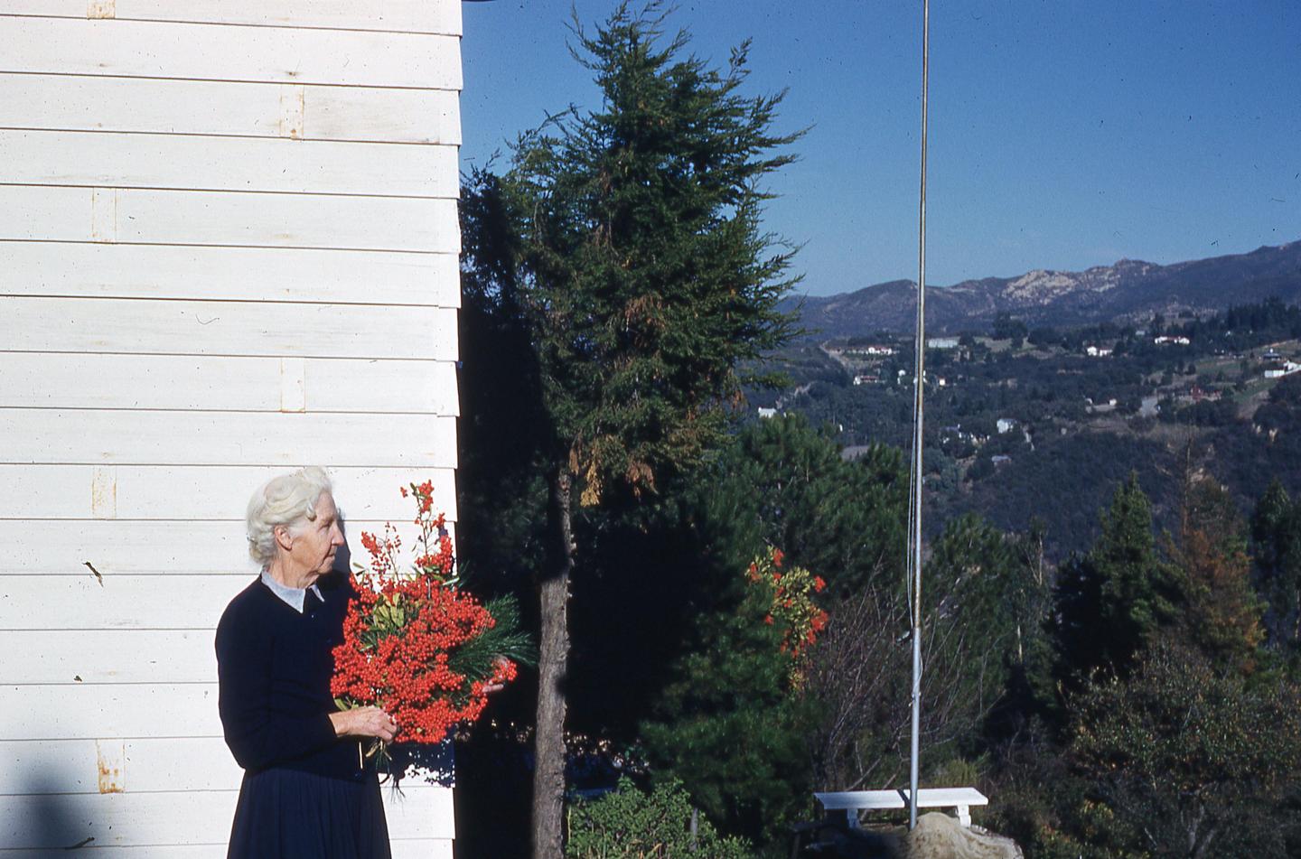 A woman stands next to a white building, holding a large bouquet of red berries and green foliage, with a mountainous landscape in the background.