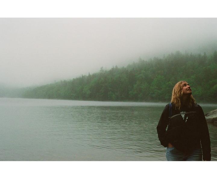 A woman stands by a lake with a forested shore shrouded in fog.