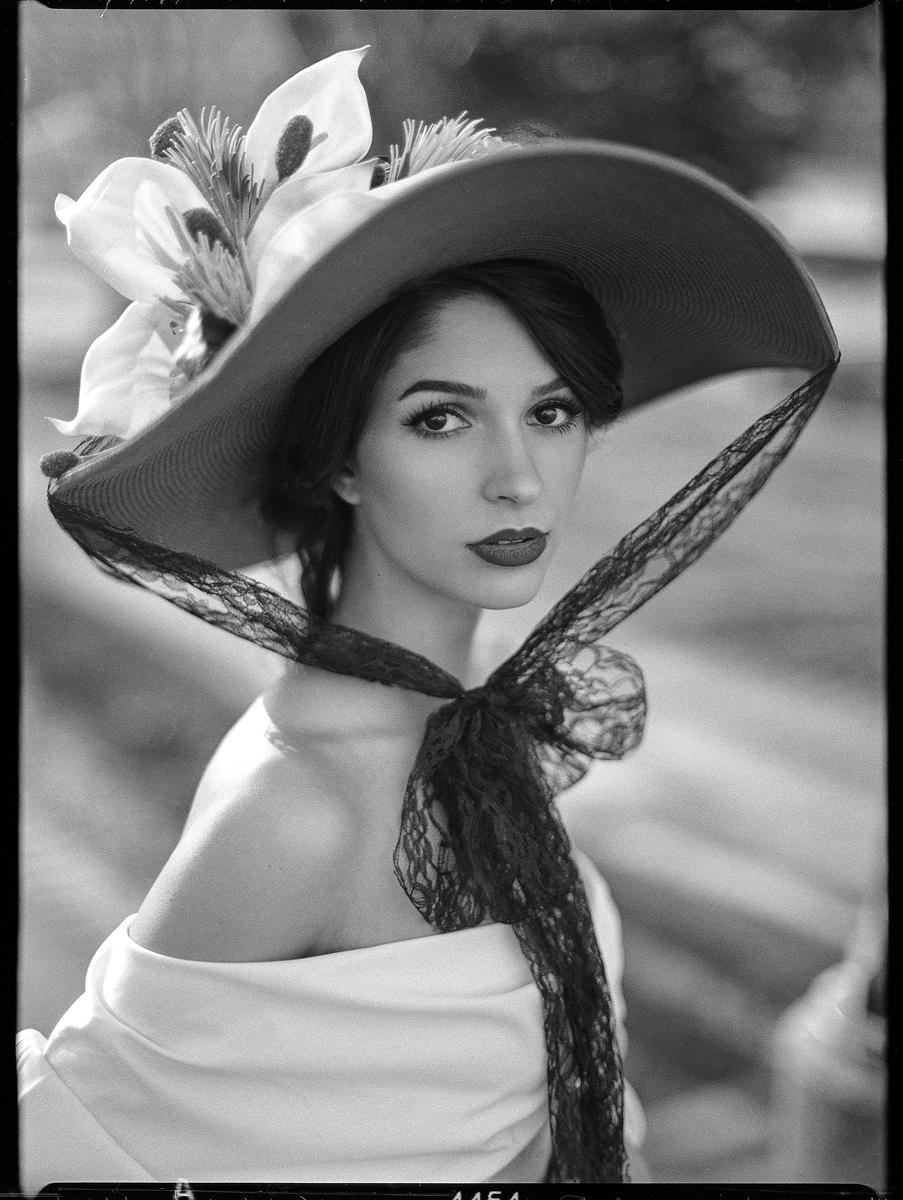 A woman wearing a wide-brimmed hat with a large flower and a lace scarf looks directly at the viewer.