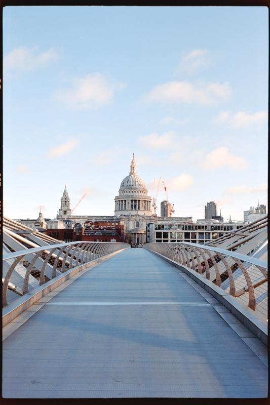 A long pedestrian bridge leads to a large domed building under a blue sky with scattered clouds.