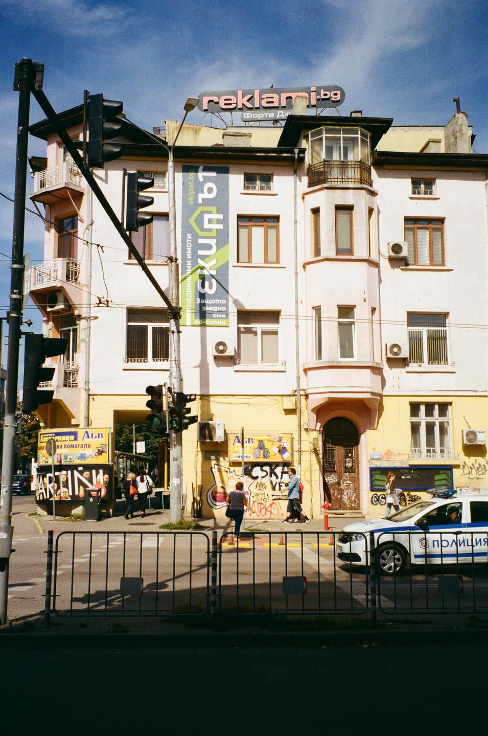 A white and yellow building with advertisements and graffiti stands at a street corner with people walking on the sidewalk and a police car parked nearby.