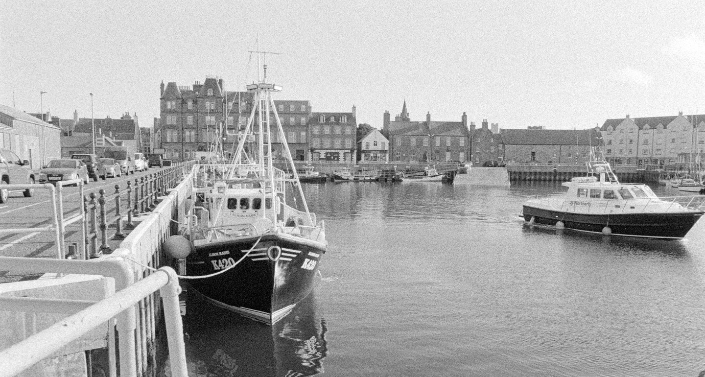 A black and white photograph shows a harbor with boats docked and buildings lining the waterfront.