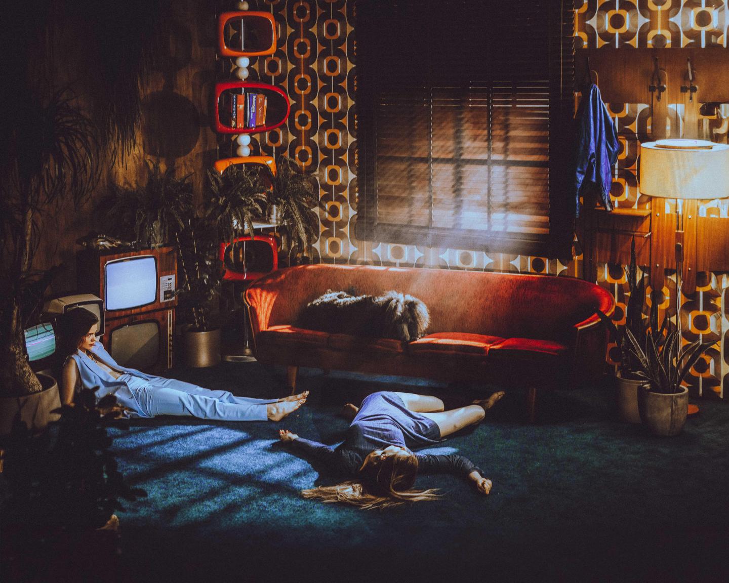 Two women are lying on a carpeted floor in a room with retro decor and plants.