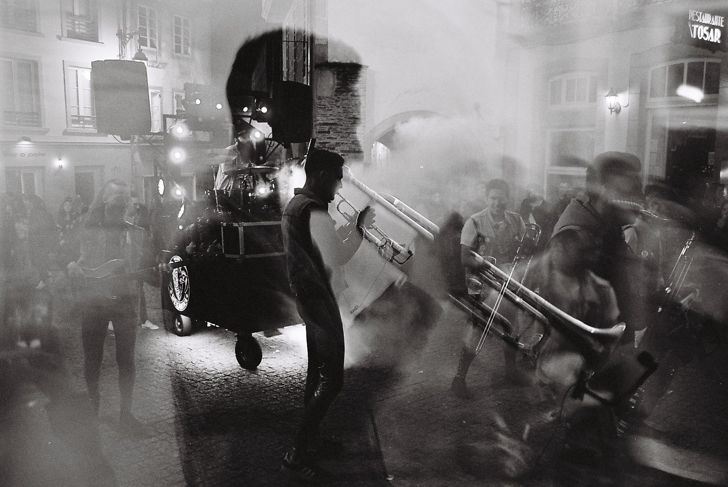 A street party with musicians playing instruments and a crowd of people is captured in a double exposure.