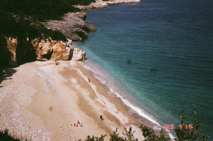 A high-angle view shows a sandy beach with a rocky cliff on the left and clear blue water with small waves on the right.