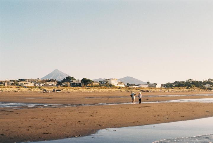 Two people walk on a wide sandy beach with houses and a mountain in the distance under a clear sky.