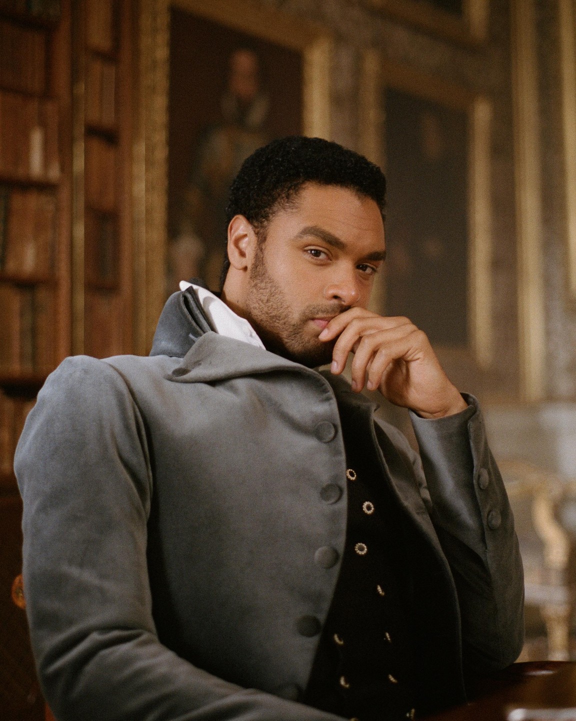 A man in period costume sits with his hand to his chin, looking directly at the viewer.
