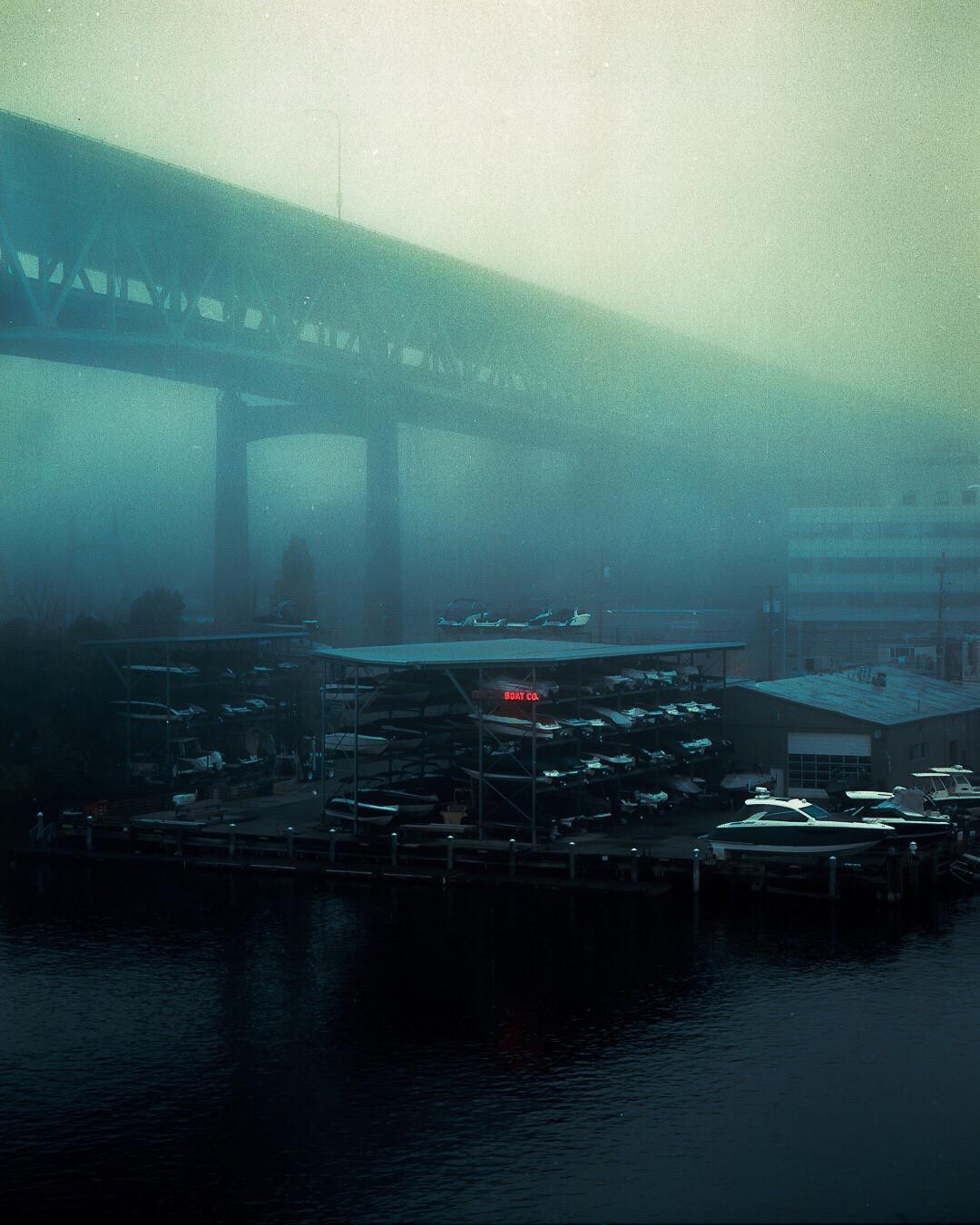 A boat storage facility with a neon sign is visible through the fog under a bridge.