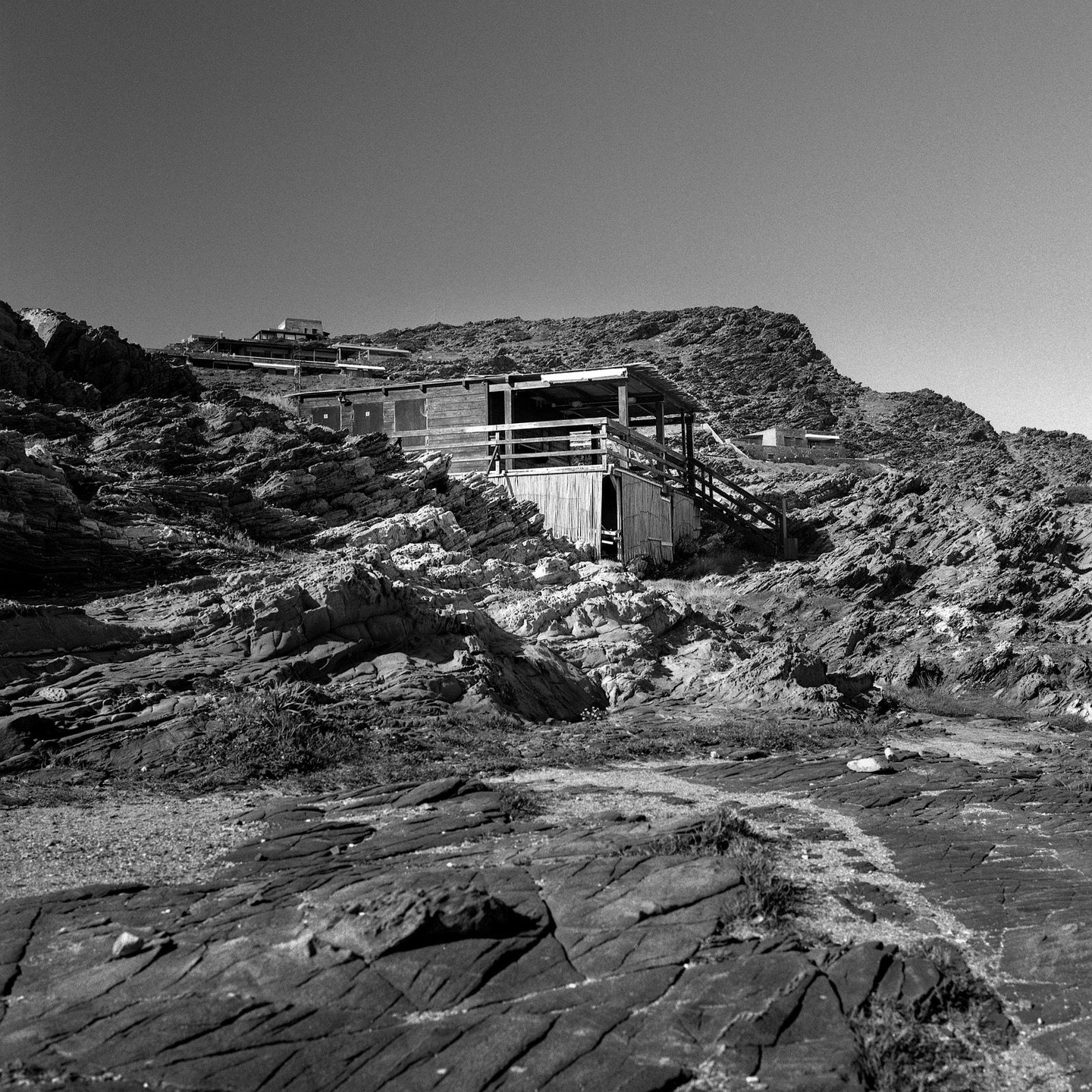 A wooden cabin with a porch and stairs is built into a rocky hillside under a clear sky.