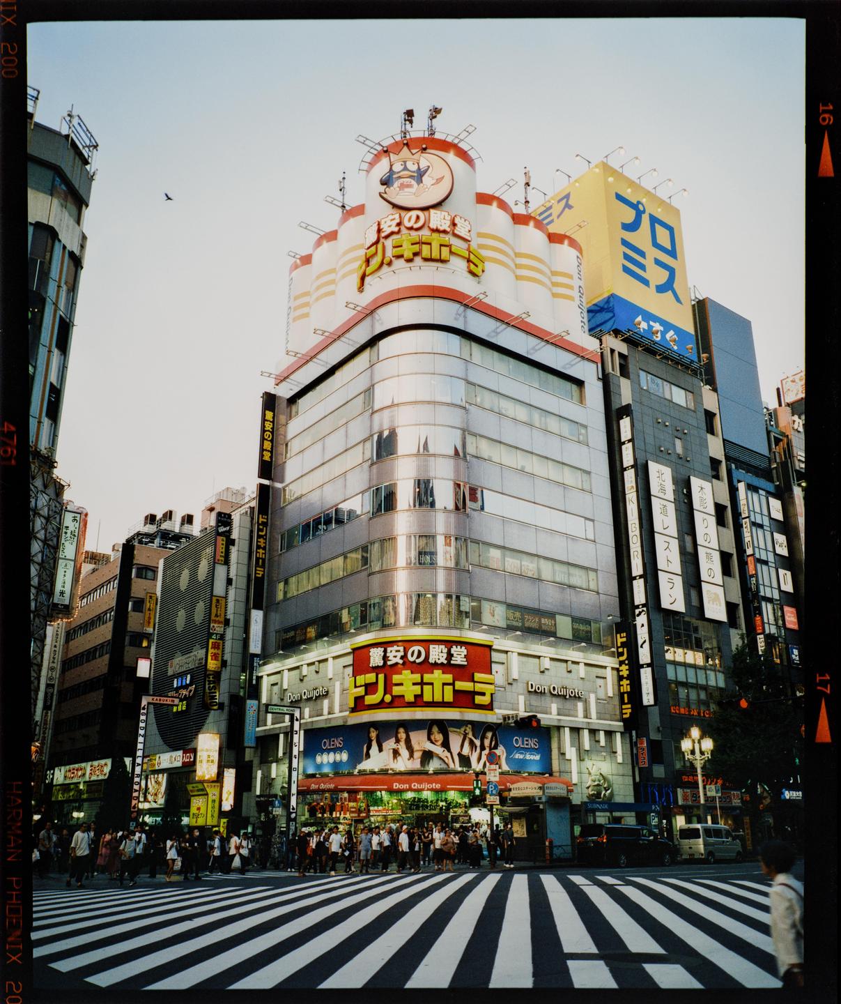 A large building with many signs and advertisements stands at a busy intersection with people crossing the street.