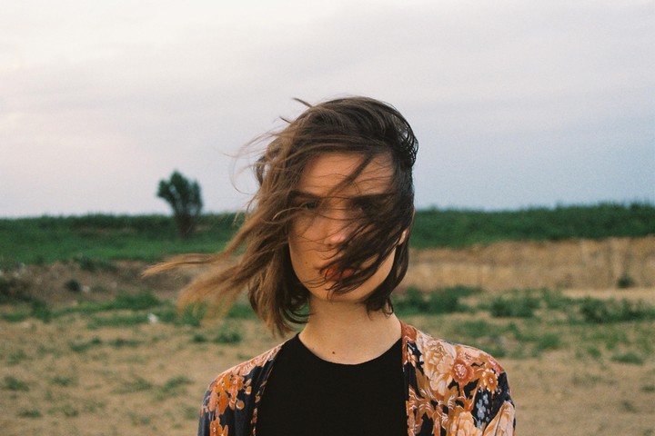 A woman with windblown hair stands in a field with a tree in the background.